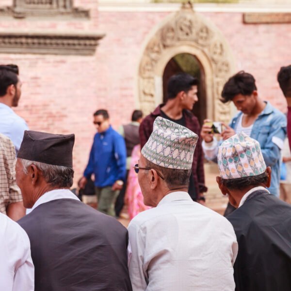 A group of men wearing traditional Nepalese Dhaka topi hats gathered in a public square, with the backs of their heads facing the camera and an ornate building entrance in the background.