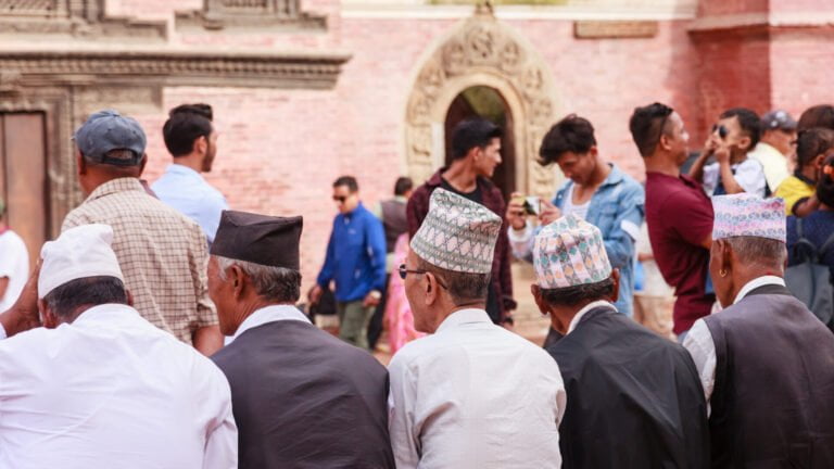 A group of men wearing traditional Nepalese Dhaka topi hats gathered in a public square, with the backs of their heads facing the camera and an ornate building entrance in the background.