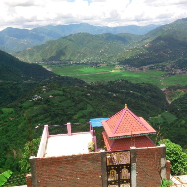 A scenic view overlooking lush green mountains with a valley and terraced fields. In the foreground, a part of a building with a red roof and ornate details is visible.