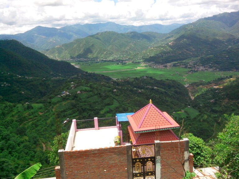 A scenic view overlooking lush green mountains with a valley and terraced fields. In the foreground, a part of a building with a red roof and ornate details is visible.