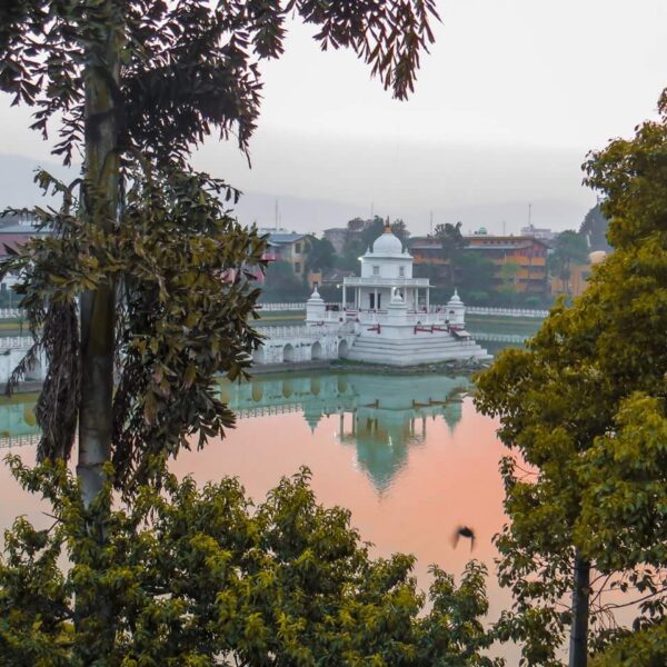 A serene view of a white temple with a domed roof, reflected in the calm waters of a pond during a misty sunrise, flanked by lush green trees with a silhouette of distant hills in the background.