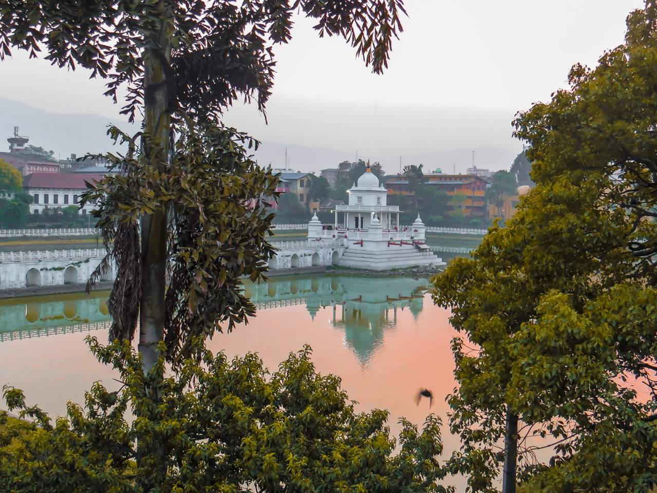 A serene view of a white temple with a domed roof, reflected in the calm waters of a pond during a misty sunrise, flanked by lush green trees with a silhouette of distant hills in the background.