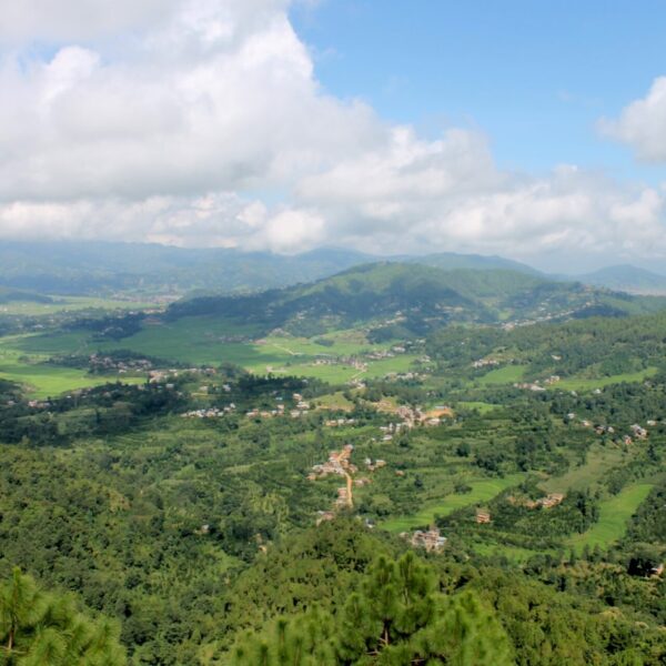 Aerial view of a lush valley with scattered settlements, surrounded by rolling green hills and covered by a mix of clouds and blue sky.