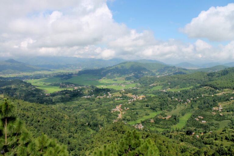 Aerial view of a lush valley with scattered settlements, surrounded by rolling green hills and covered by a mix of clouds and blue sky.