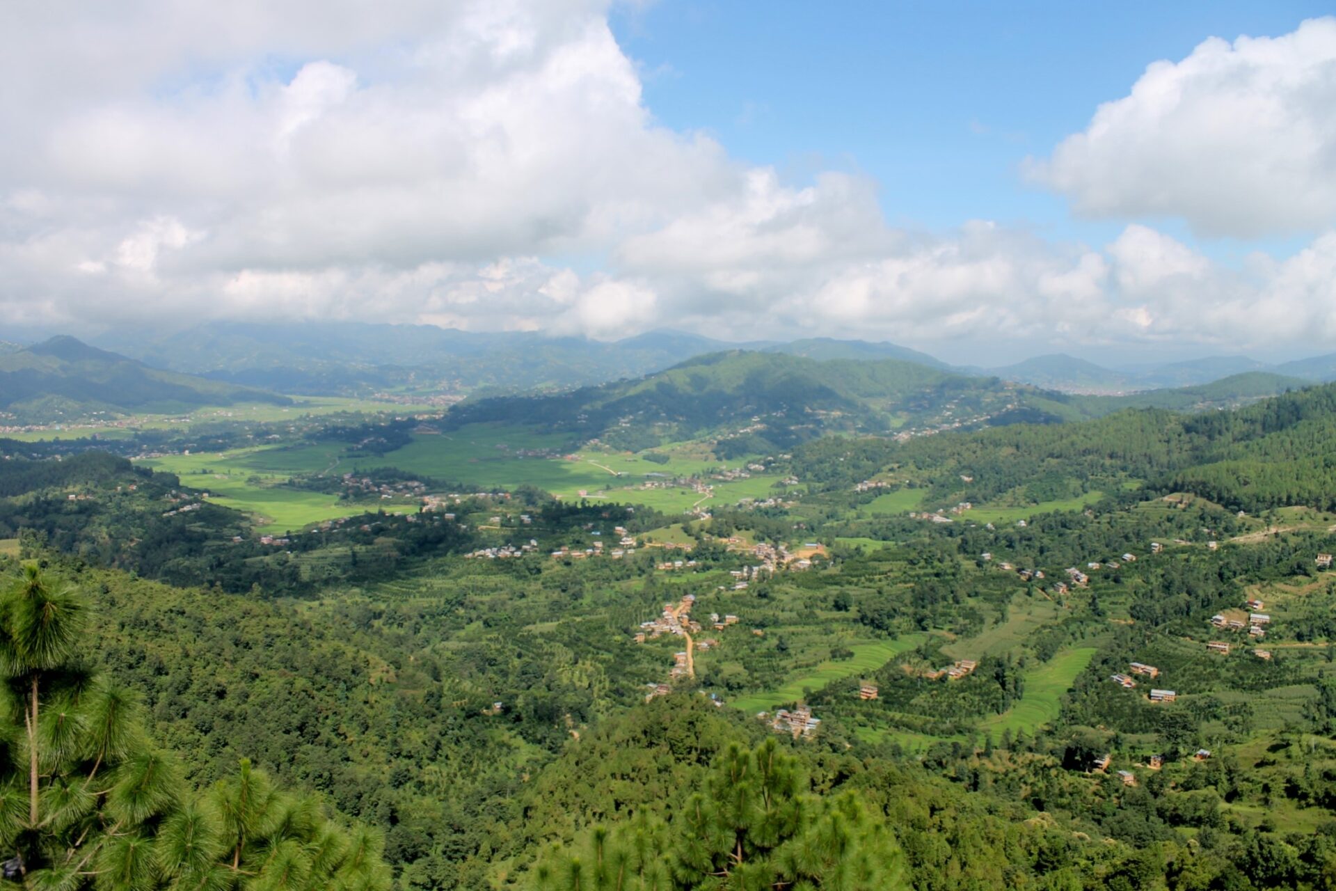 Aerial view of a lush valley with scattered settlements, surrounded by rolling green hills and covered by a mix of clouds and blue sky.