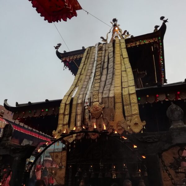 Alt text: A traditional Nepalese chariot with ornate decorations and fabric coverings, part of a festival, displaying intricate wood carvings and colorful details, against a backdrop of a temple and a clear sky.