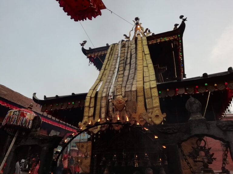 Alt text: A traditional Nepalese chariot with ornate decorations and fabric coverings, part of a festival, displaying intricate wood carvings and colorful details, against a backdrop of a temple and a clear sky.