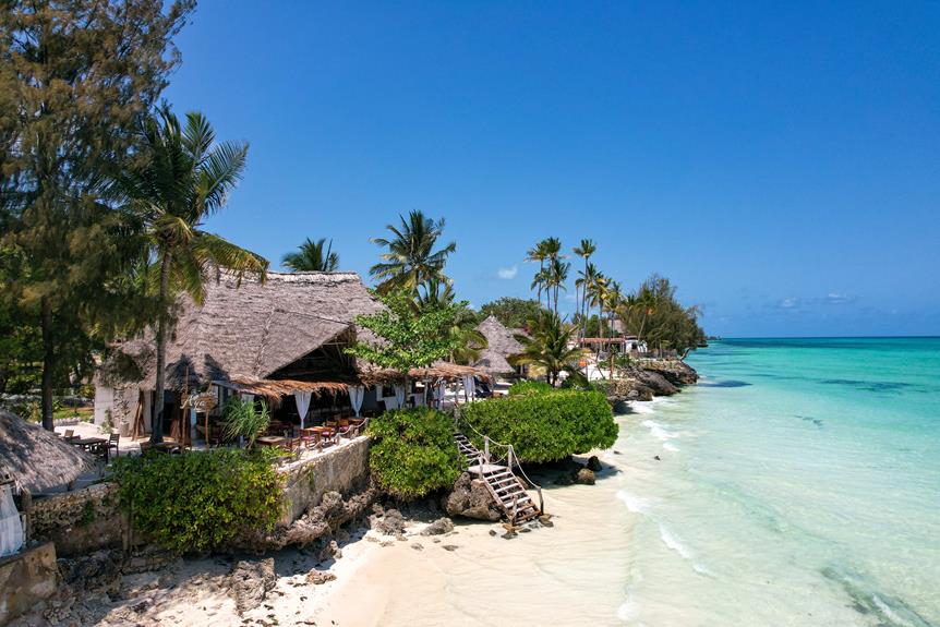 A tropical beachside resort with thatched-roof structures, palm trees, clear turquoise waters, and a small sandy beach.