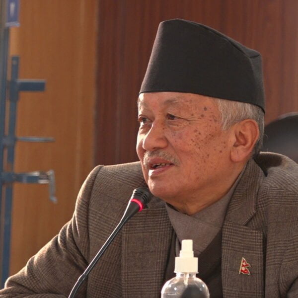 An older man wearing a traditional Nepali Dhaka topi and a suit sits before a microphone, appearing to be in a conference or meeting room.