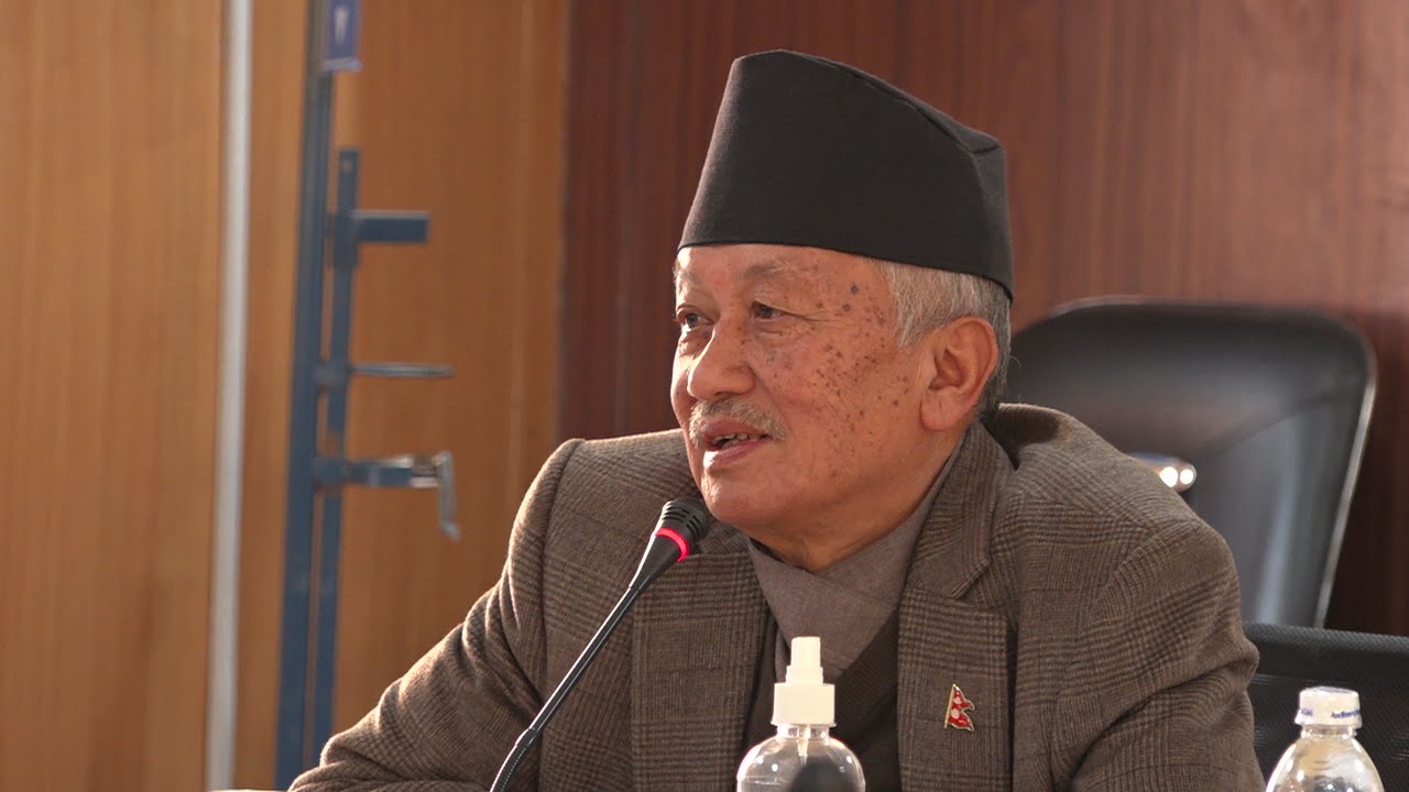 An older man wearing a traditional Nepali Dhaka topi and a suit sits before a microphone, appearing to be in a conference or meeting room.