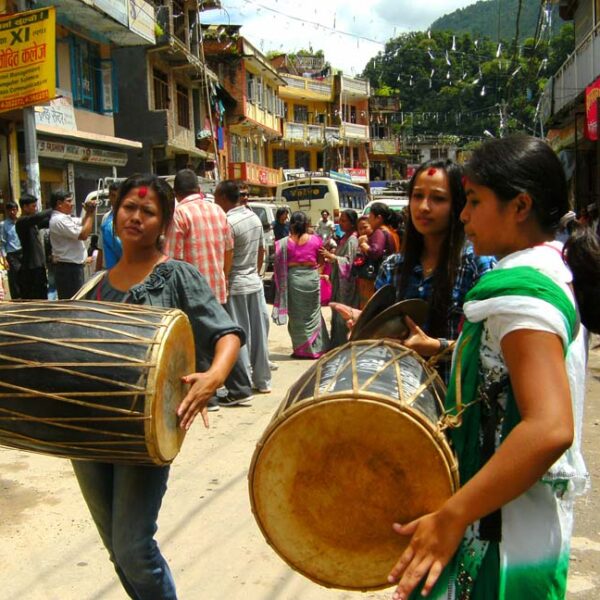 Three women carrying traditional drums on a bustling street in a mountainous town with people and shops in the background.