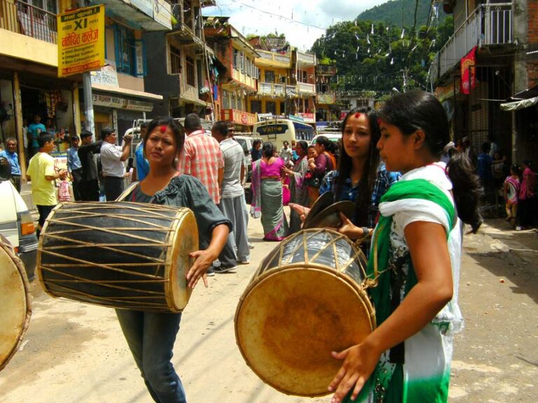 Three women carrying traditional drums on a bustling street in a mountainous town with people and shops in the background.