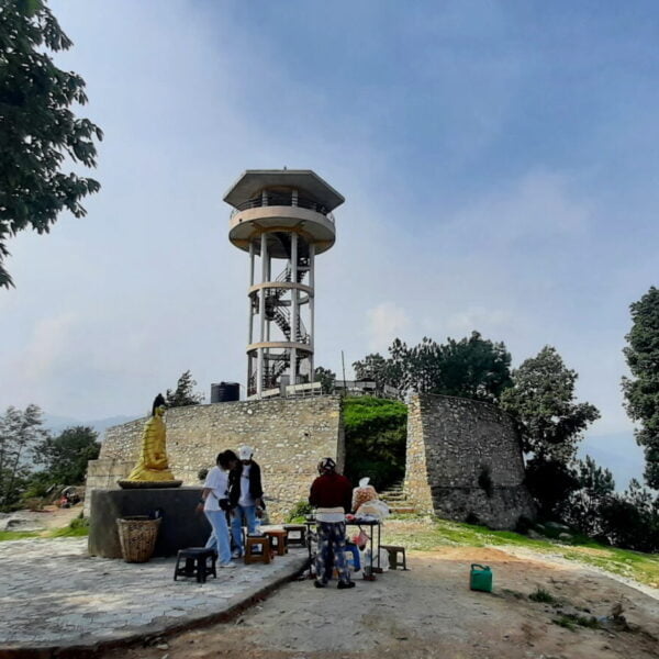 A group of people gathered around a concrete viewing tower with a small Buddha statue in front, surrounded by greenery and with a hazy blue sky above.