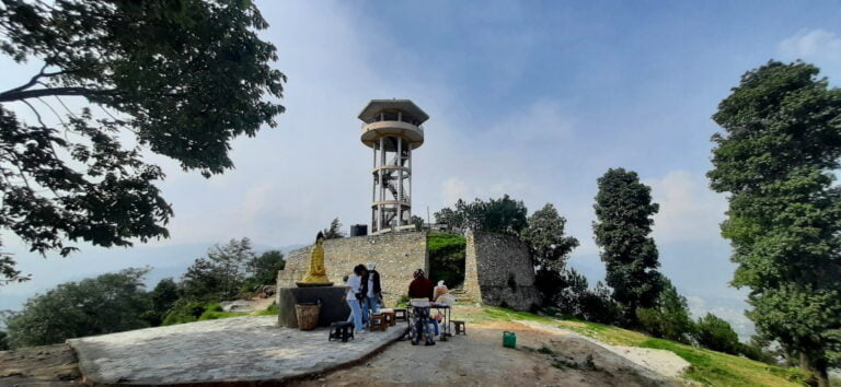 A group of people gathered around a concrete viewing tower with a small Buddha statue in front, surrounded by greenery and with a hazy blue sky above.