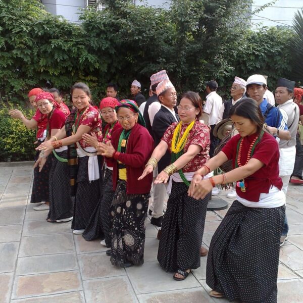 A group of people in traditional Nepalese attire performing a dance outdoors, with some spectators in the background.