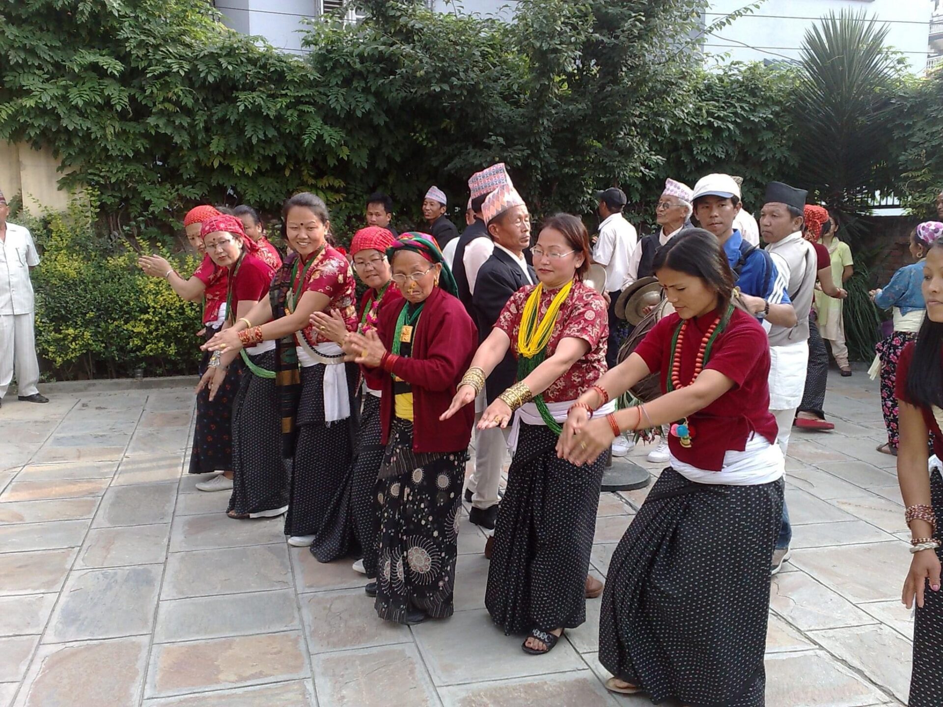 A group of people in traditional Nepalese attire performing a dance outdoors, with some spectators in the background.