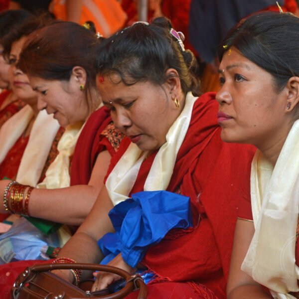 A group of women in traditional red and gold attire with shawls and decorative bindis sitting side by side at an event.