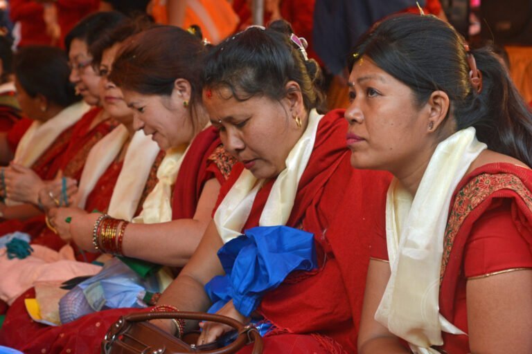 A group of women in traditional red and gold attire with shawls and decorative bindis sitting side by side at an event.