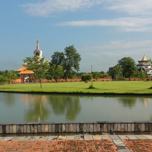 A picturesque view of a calm lake reflecting a serene landscape with traditional Asian-style buildings and a pagoda, green trees, clear skies, and a brick pathway in the foreground.
