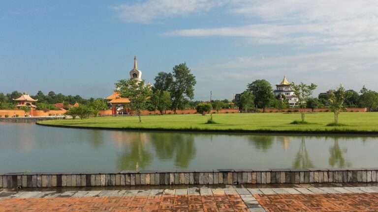 A picturesque view of a calm lake reflecting a serene landscape with traditional Asian-style buildings and a pagoda, green trees, clear skies, and a brick pathway in the foreground.
