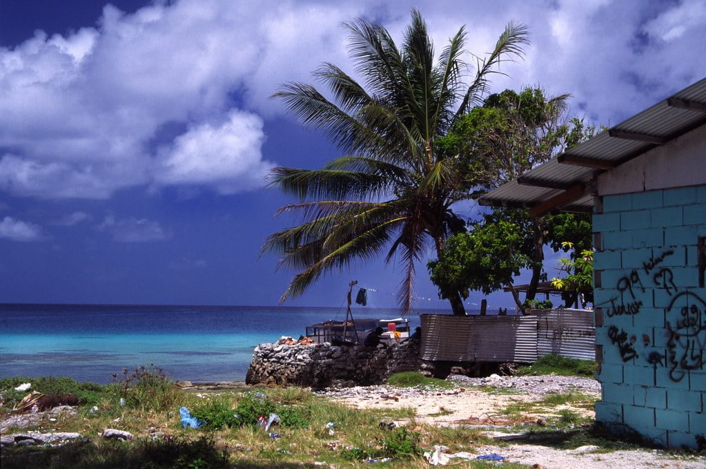 A coastal scene with a clear turquoise sea under a blue sky with fluffy clouds. A palm tree leans over a littered beach next to a building with graffiti.