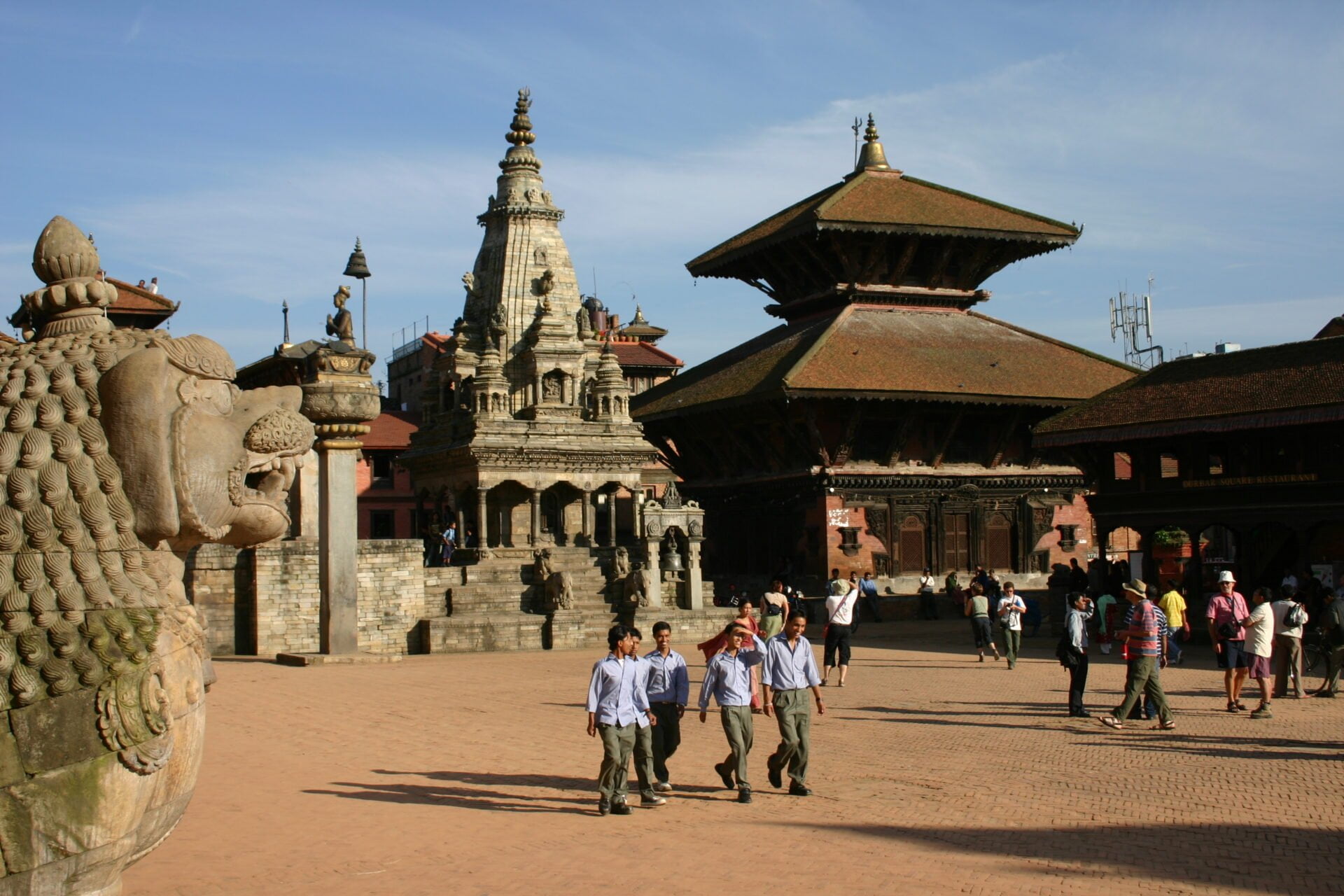 Alt text: People walking and socializing on a spacious courtyard surrounded by intricate Nepalese-style temples and historic architecture under a clear blue sky. A stone statue is visible on the left.