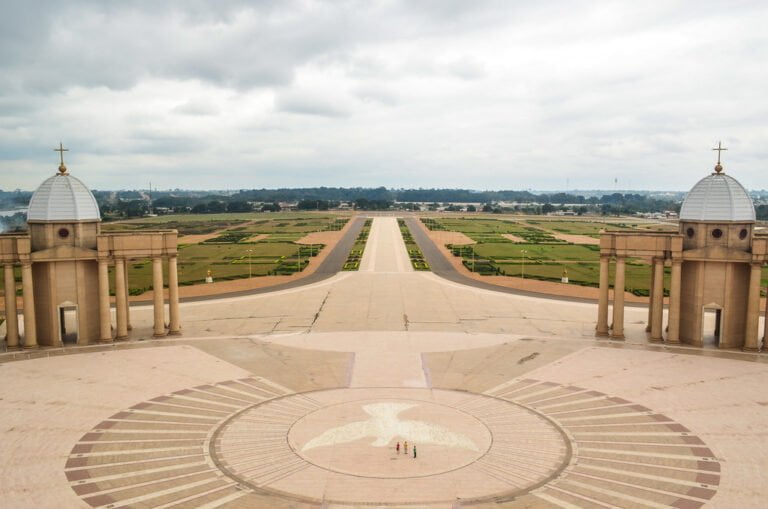 A view from above showing a large open plaza with a radial pattern on the ground, flanked by two symmetrical buildings with domes, overlooking a vast landscaped area with fields and trees in the distance under a cloudy sky.
