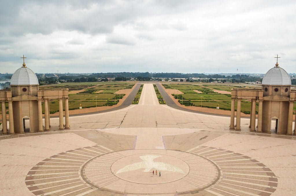A view from above showing a large open plaza with a radial pattern on the ground, flanked by two symmetrical buildings with domes, overlooking a vast landscaped area with fields and trees in the distance under a cloudy sky.