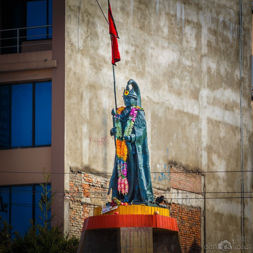 A colorful statue of a figure with a flagpole stands adorned with fresh flower garlands against a backdrop of a plain building wall.