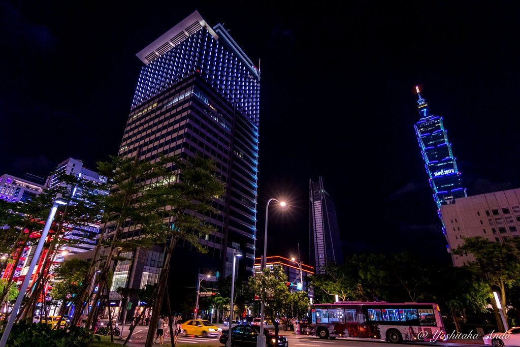 A nighttime cityscape with illuminated buildings, including a prominent skyscraper with blue lighting, traffic on the street with motion blur on vehicles, and lit trees in the foreground.