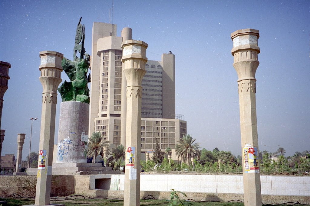 A photo depicting a public square with tall columns, sculptures, and modern buildings in the background, alongside greenery and a clear sky.