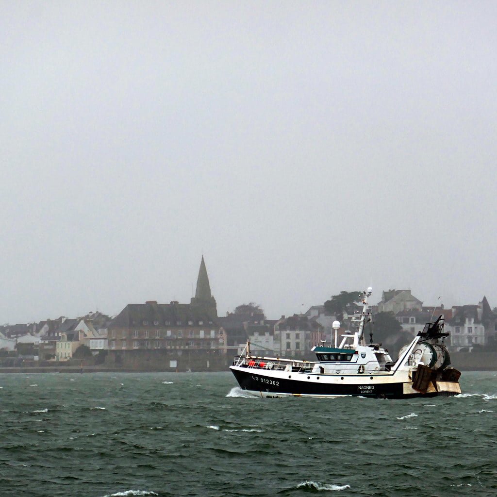 A fishing boat sailing on choppy waters with a coastal town and a church spire in the background on an overcast day.