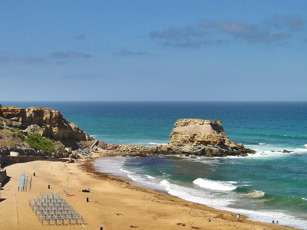 A scenic beach with golden sand, surrounded by rocky cliffs against a blue sky, with the ocean waves crashing onto the shore.