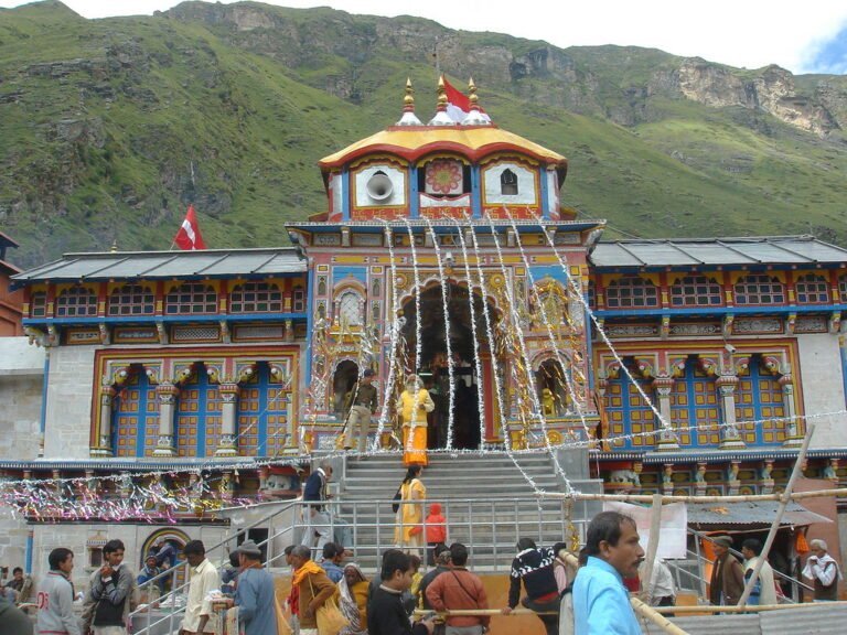A vibrant and colorful temple with traditional architecture nestled against a backdrop of green mountains, with people visiting and socializing in the foreground.