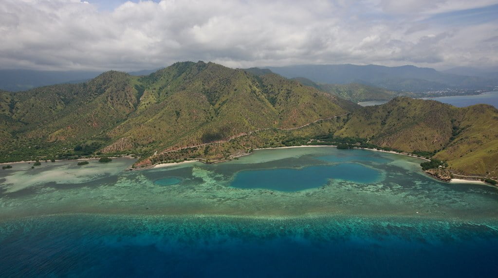Aerial view of a tropical coastline with mountains, showing clear turquoise waters with coral reefs and a partly cloudy sky.