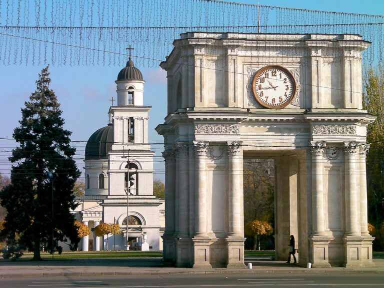 Stone archway with a clock in the foreground and a church with a dome and bell tower in the background, under a clear blue sky. A person is visible walking by the archway.