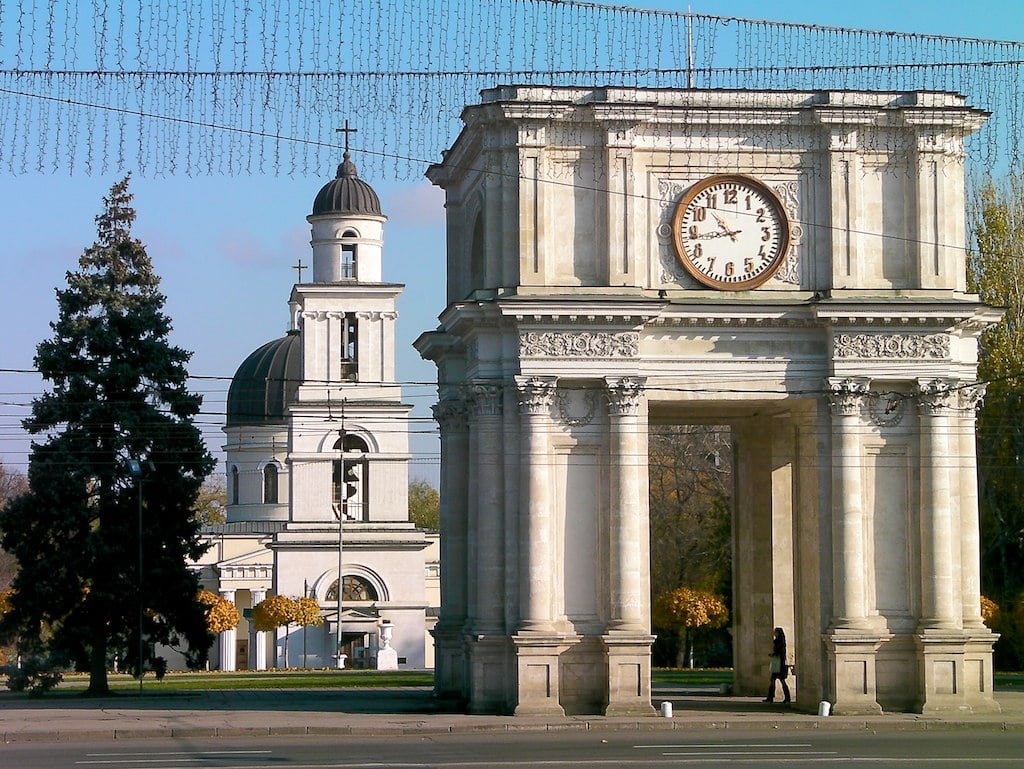 Stone archway with a clock in the foreground and a church with a dome and bell tower in the background, under a clear blue sky. A person is visible walking by the archway.