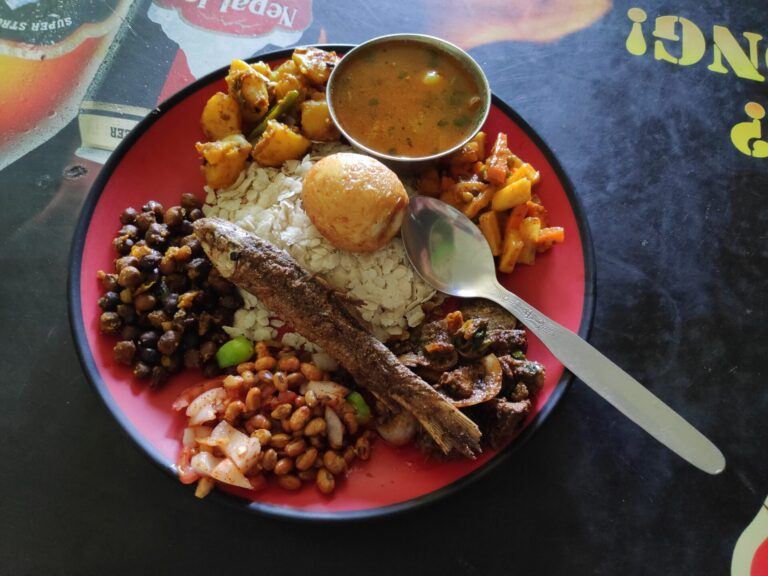 A plate of assorted dishes including rice, a grilled fish, beans, potatoes, salad, a bread roll and a bowl of soup, with a spoon on the side, on a tabletop with a partially visible logo.