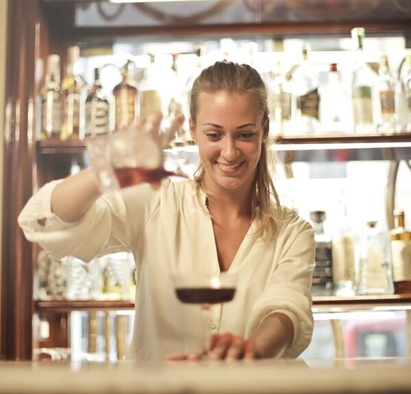 A smiling female bartender in a white blouse serving a red cocktail at a bar with a variety of bottles in the background.