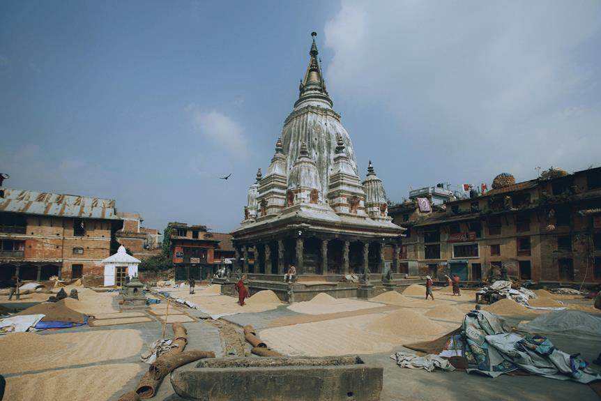 An ancient, weathered temple with a tall, ornate spire surrounded by heaps of grain drying in the sun, with people walking by and traditional buildings in the backdrop under a clear sky.