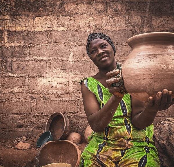 A woman in a green printed dress sitting in front of a brick wall while holding and examining a handmade clay pot, with other pottery pieces nearby.