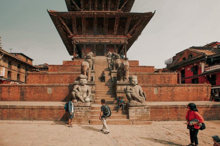 Stone statues of mythical creatures flank the steps leading up to a traditional tiered temple in a square with people milling around.