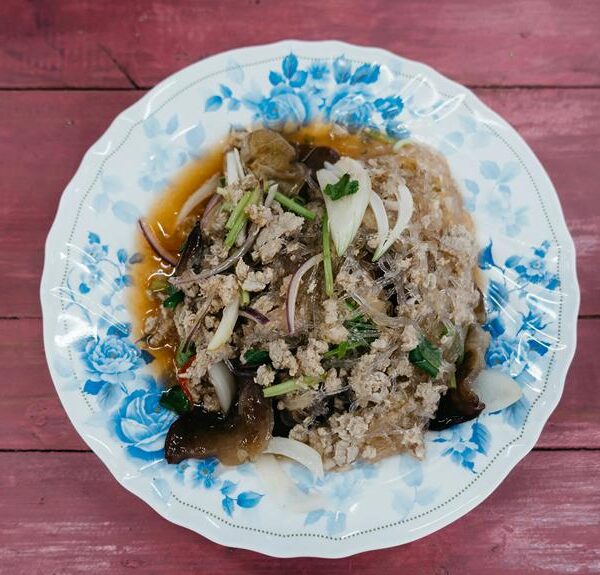 A traditional Asian dish with minced meat, glass noodles, and vegetables on a floral-patterned plate, set against a pink wooden background.