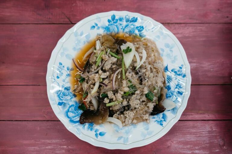 A traditional Asian dish with minced meat, glass noodles, and vegetables on a floral-patterned plate, set against a pink wooden background.