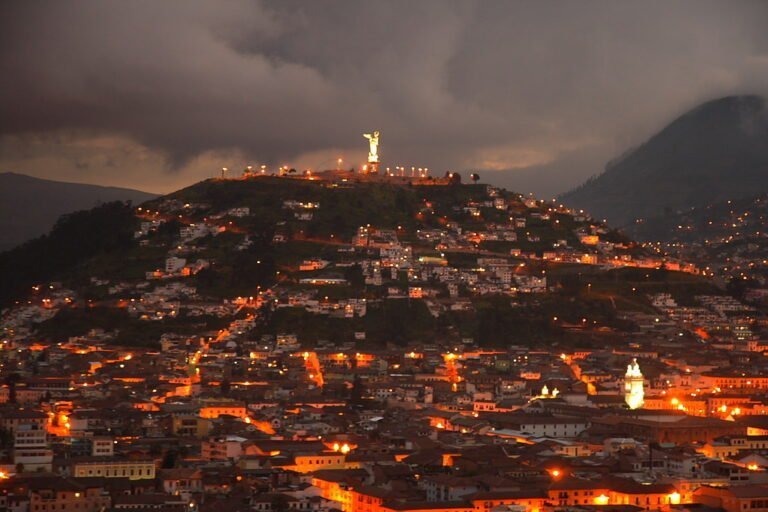 A panoramic evening view of a city with lights on, nestled on hilly terrain beneath a dark sky with a large illuminated statue atop the hill in the background.