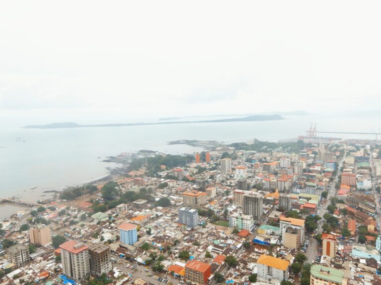 Aerial view of a densely built coastal city with various buildings, roads, and a cloudy sky, with the ocean and a distant island visible in the background.