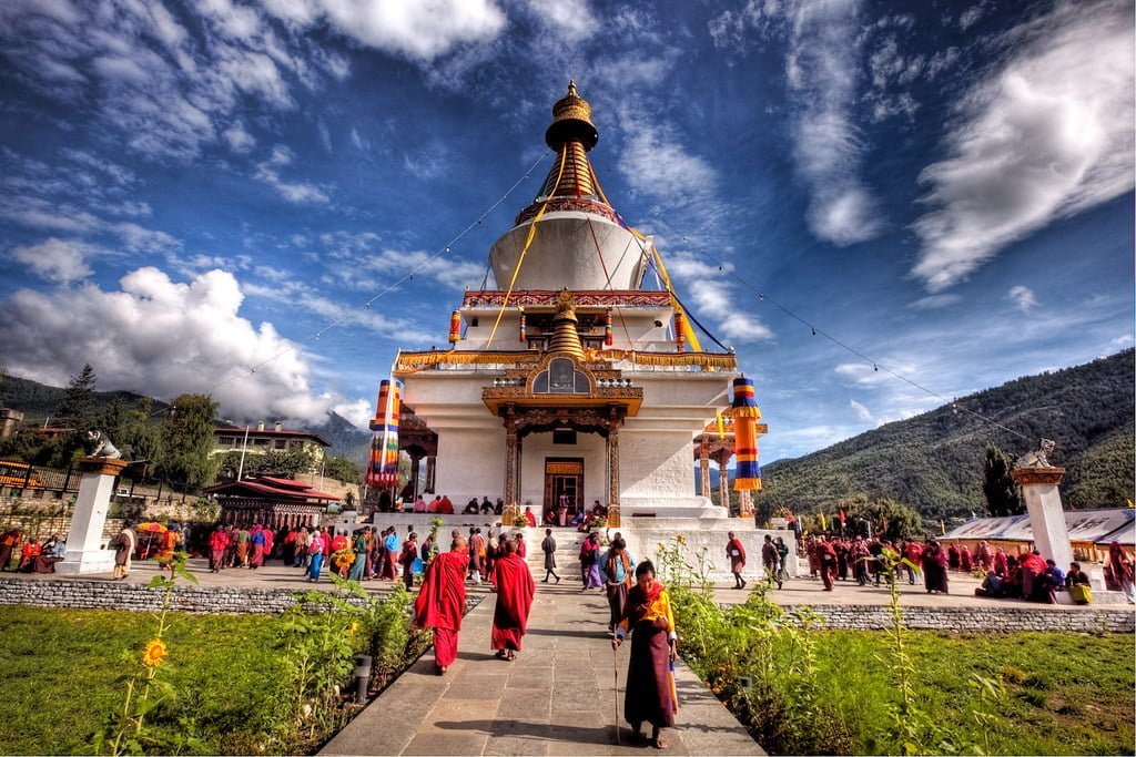 Alt text: A vibrant scene at a Buddhist monastery with a stupa in the center, adorned with colorful flags. Monks in red robes and visitors in various attire walk on the path leading to the stupa. The backdrop features a vivid blue sky with scattered clouds and lush green mountains.