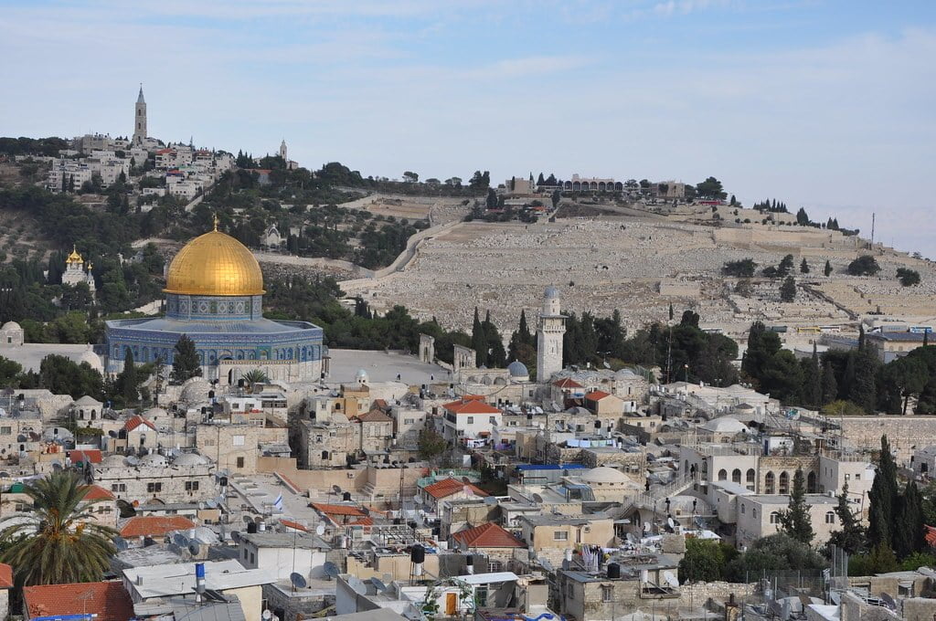 A panoramic view of Jerusalem featuring the Dome of the Rock in the foreground, surrounded by densely packed buildings, with the Mount of Olives and its cemetery in the background under a blue sky.