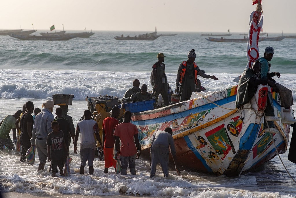 A group of people is working around a colorful wooden boat on the shore as waves crash in, with several other boats visible in the background on the water.