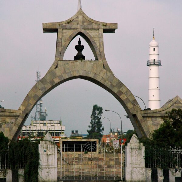 A photo showing a unique archway with a silhouetted monument at the center, adjacent to a white lighthouse against an overcast sky, with urban surroundings and a billboard to the left.